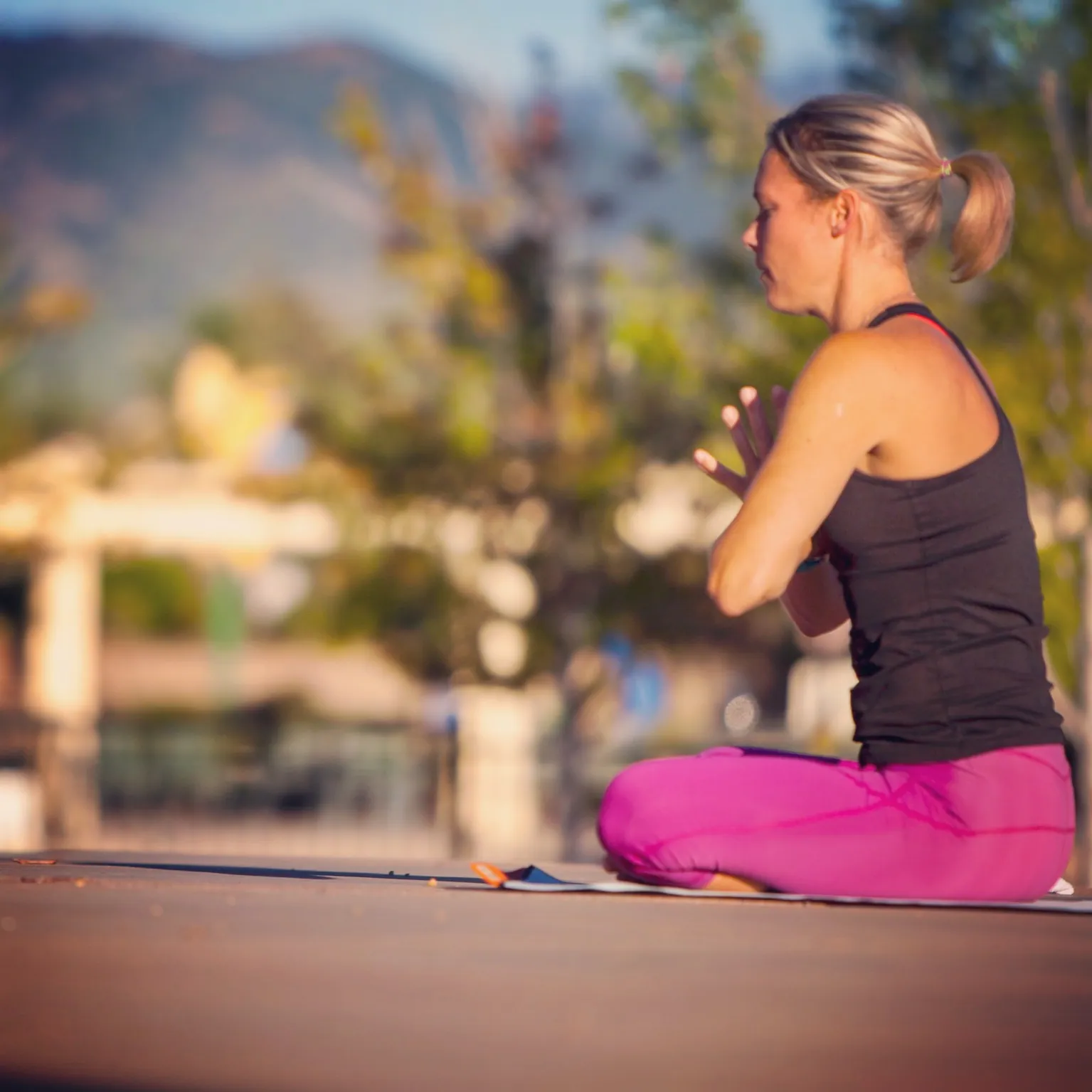 Woman sits cross-legged with hands at her heart in front of a blurred park and mountains