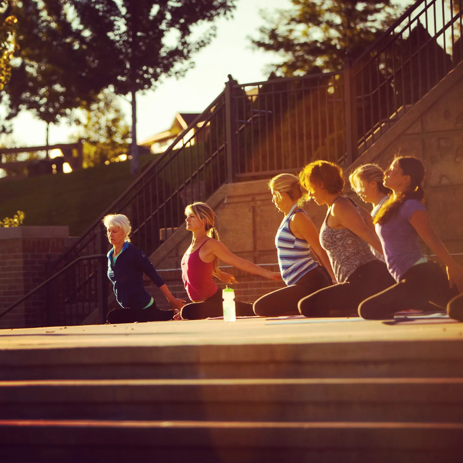 Women sit in a row on stone steps in a park during a yoga class
