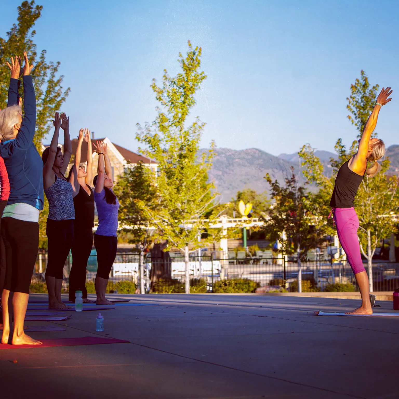 Outdoor yoga class stands with arms raised in a park with mountains behind
