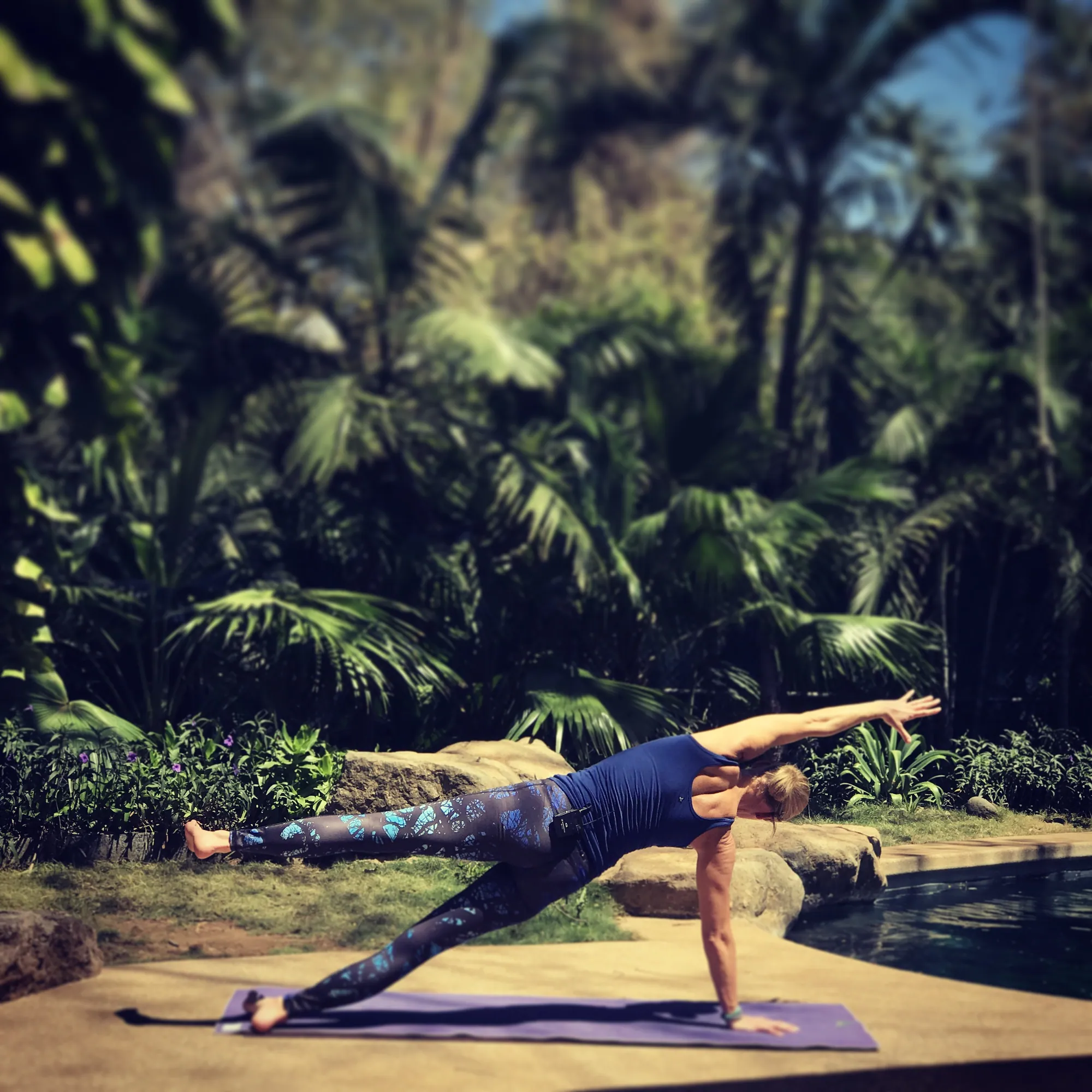 Woman in side plank on a yoga mat beside a pool surrounded by palms