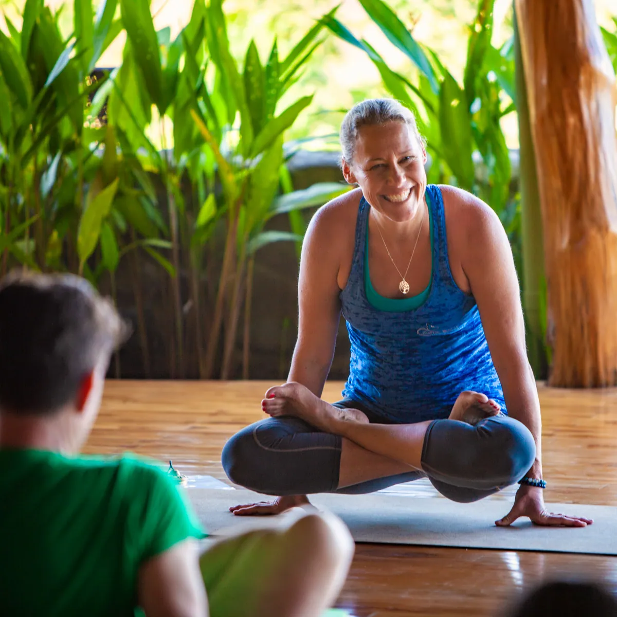 Smiling woman in a seated cross-legged balance pose surrounded by tropical plants