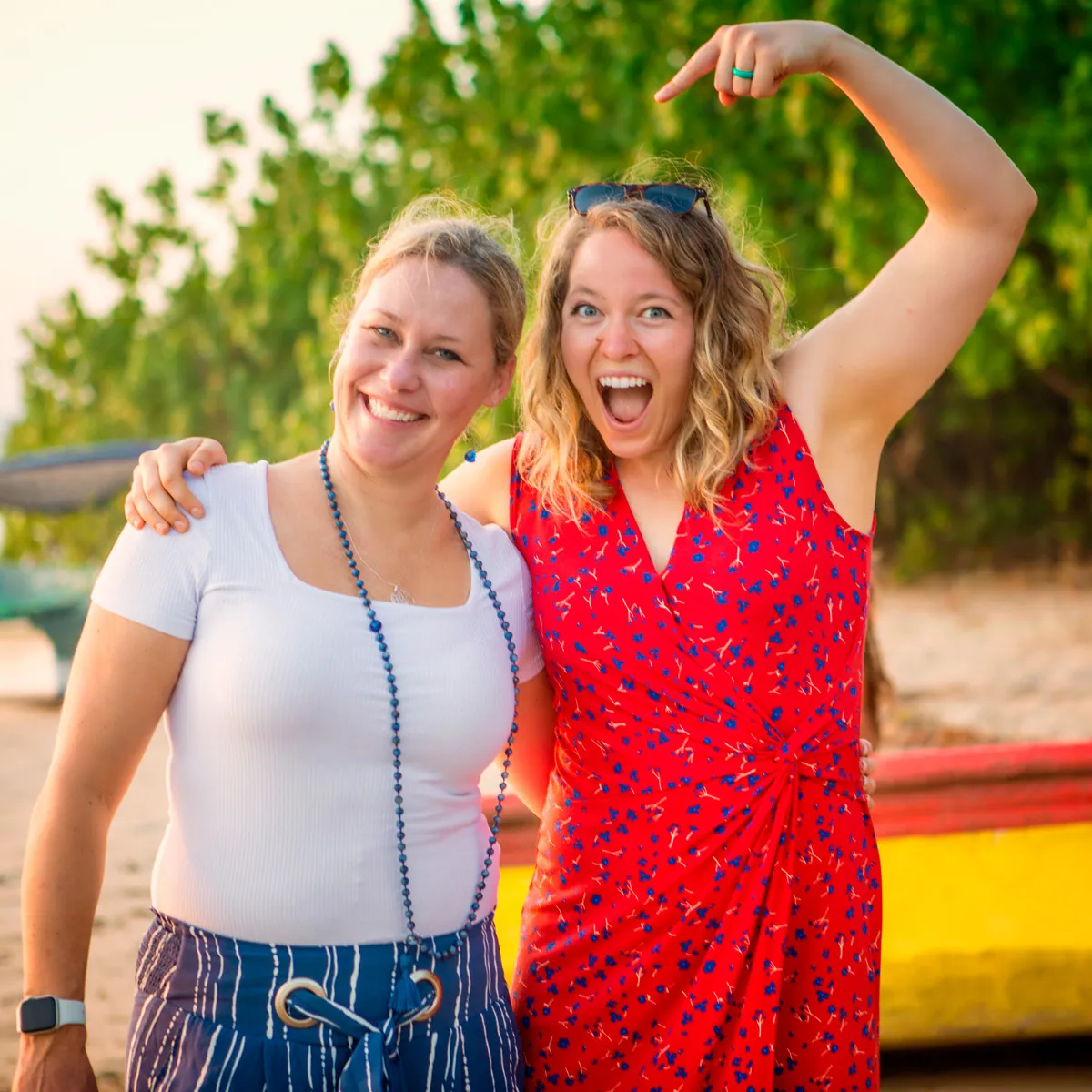 Two women laughing on a beach, one in a red dress posing with a flexed arm