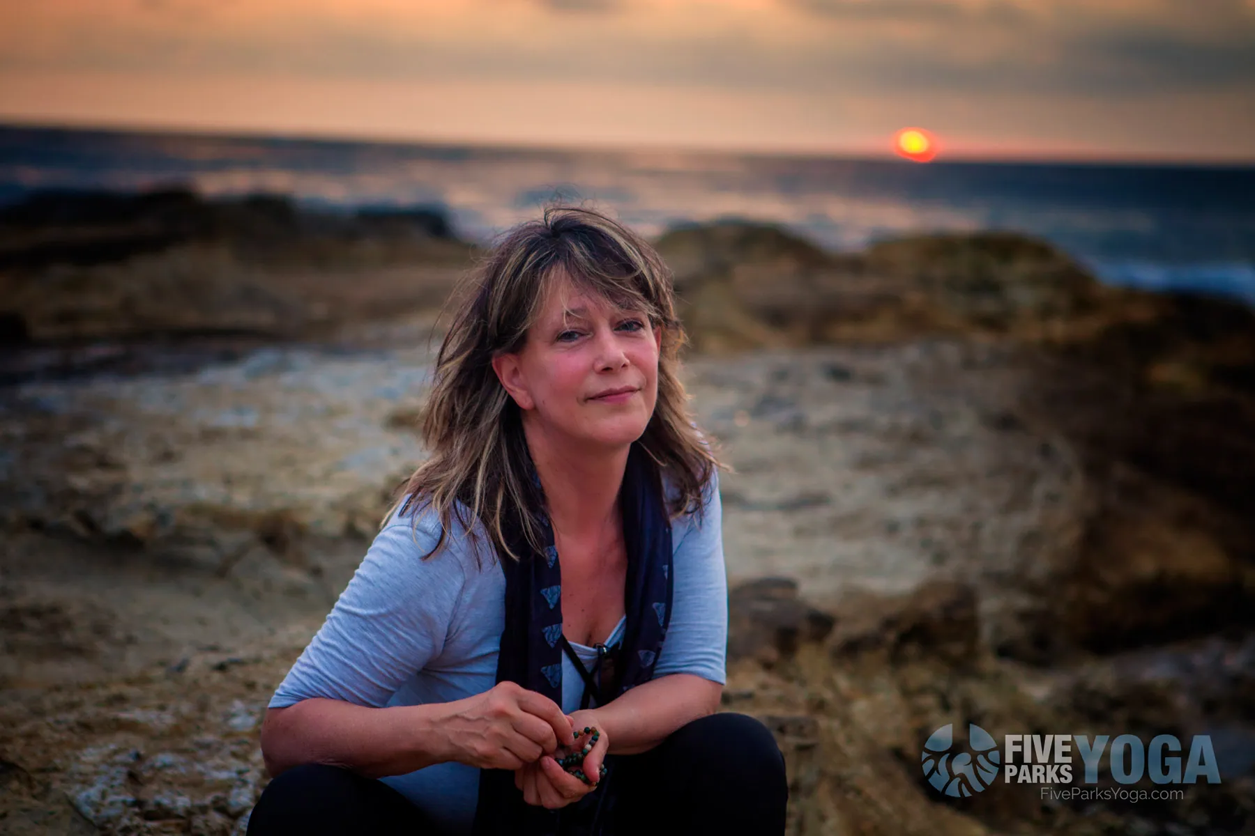 Retreat guest crouching on rocky shoreline at sunset with the ocean behind her