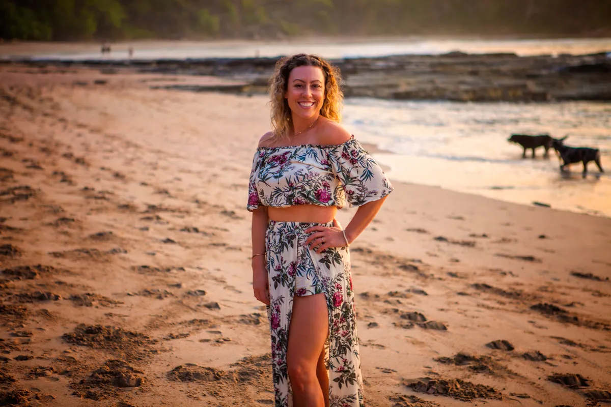 Smiling retreat guest in a floral outfit on the beach at sunset with two dogs behind her
