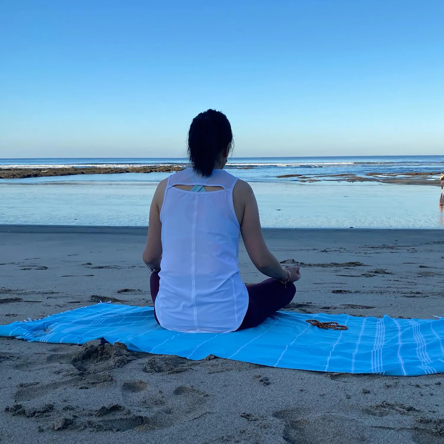 Woman meditating on a blue towel facing the ocean at the beach