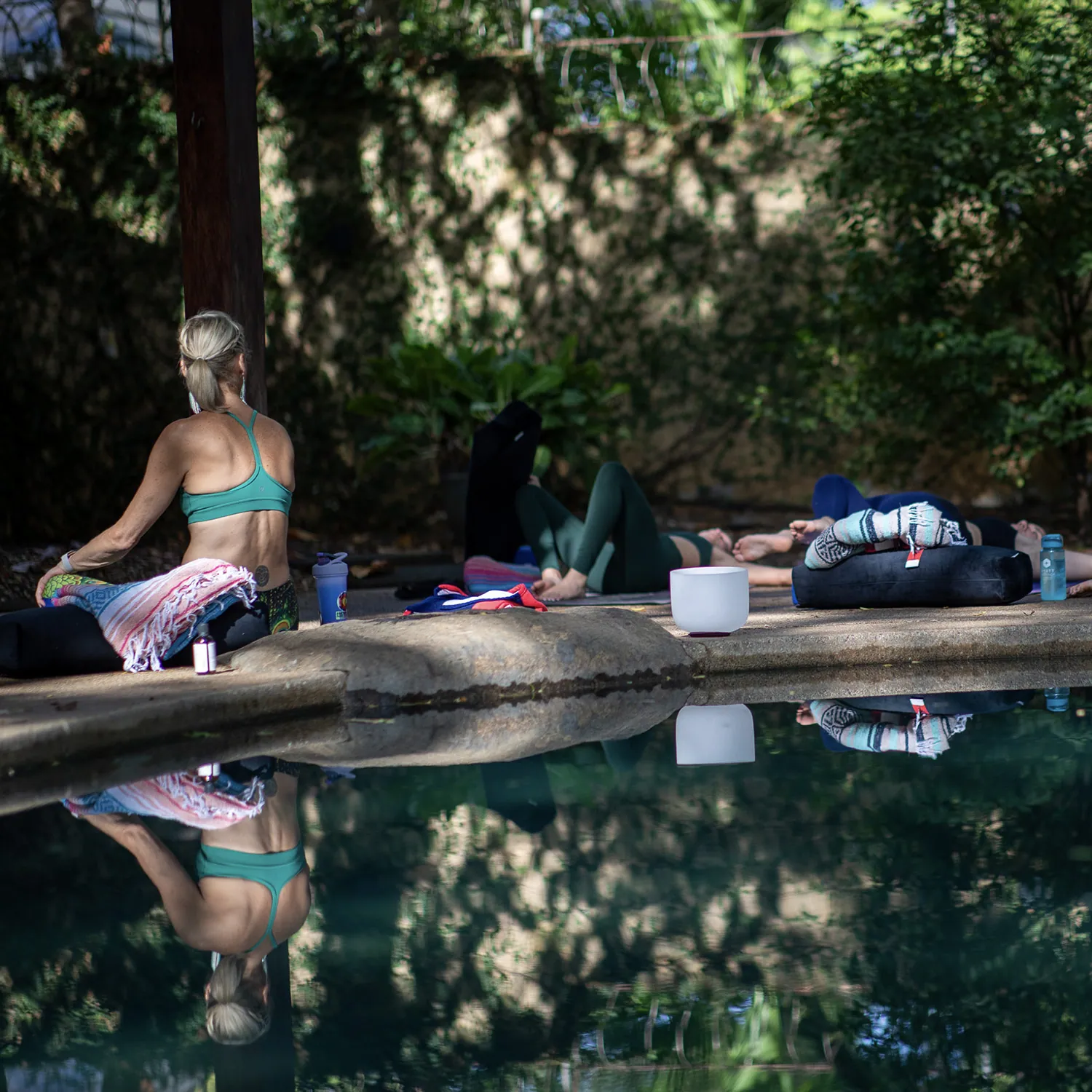 Woman meditating beside a jungle pool with a singing bowl and reflection