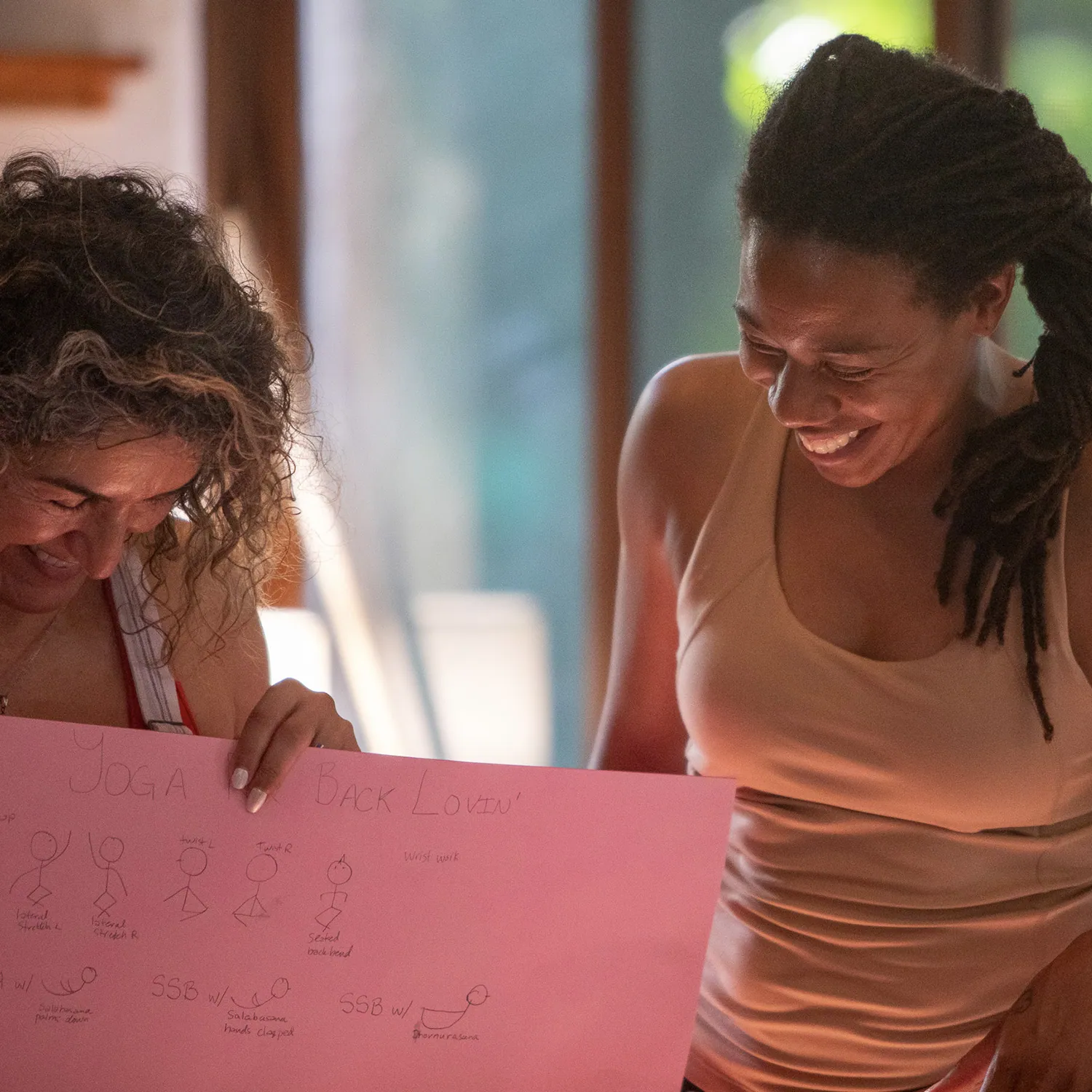 Two women laughing while holding a pink poster of yoga sketches